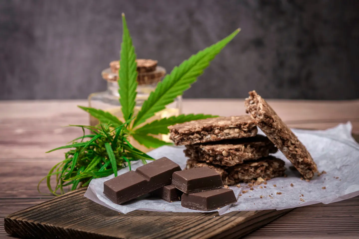 A rustic arrangement of cannabis-infused edibles, including chocolate pieces and granola bars, with fresh cannabis leaves and a jar of oil in the background.